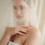 Woman wearing a white wedding dress with a bow-embellished veil against a neutral background