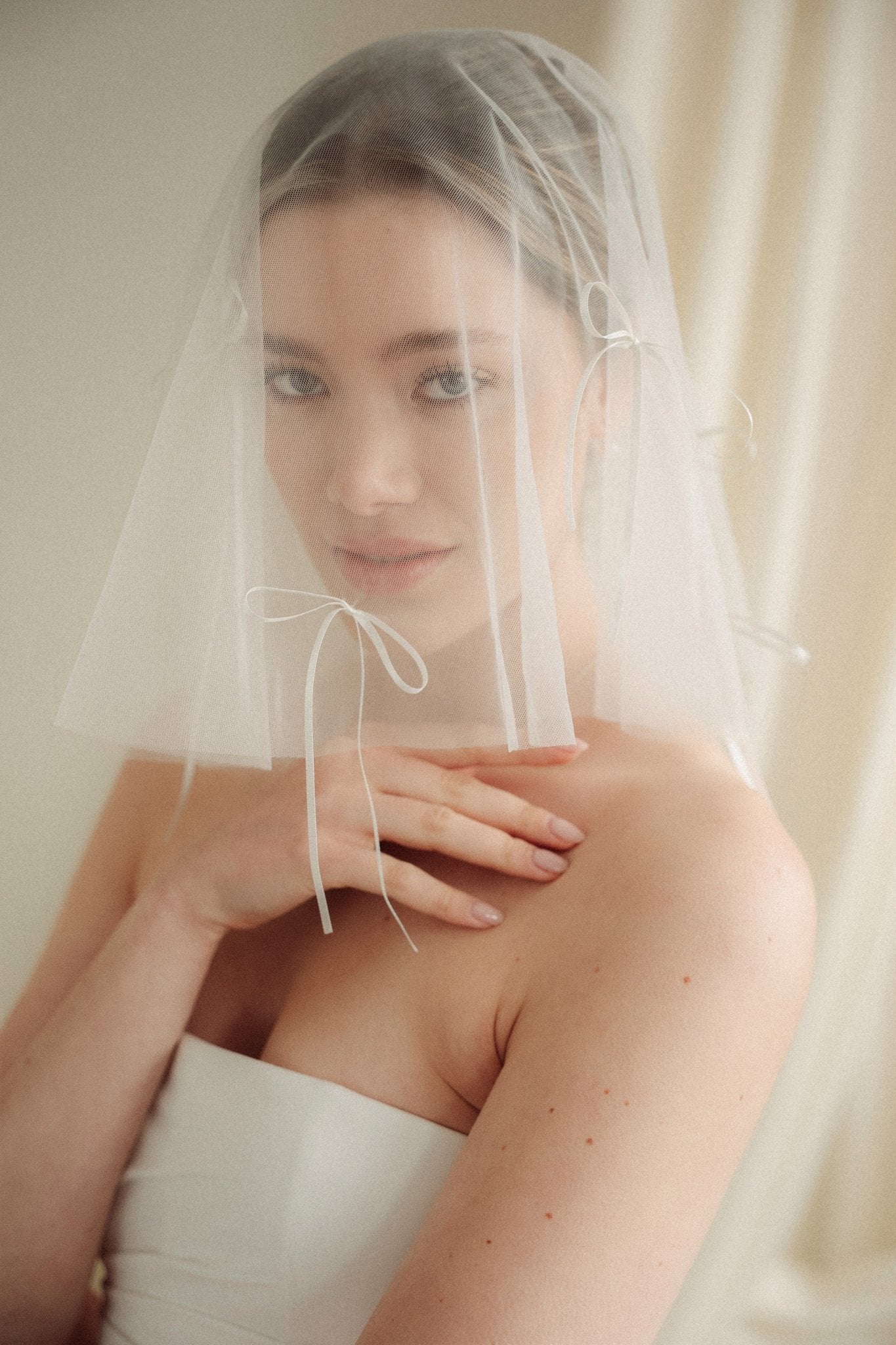 Woman wearing a white wedding dress with a bow-embellished veil against a neutral background