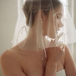 Woman wearing a white wedding dress with a bow-embellished veil against a neutral background