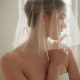 Woman wearing a white wedding dress with a bow-embellished veil against a neutral background