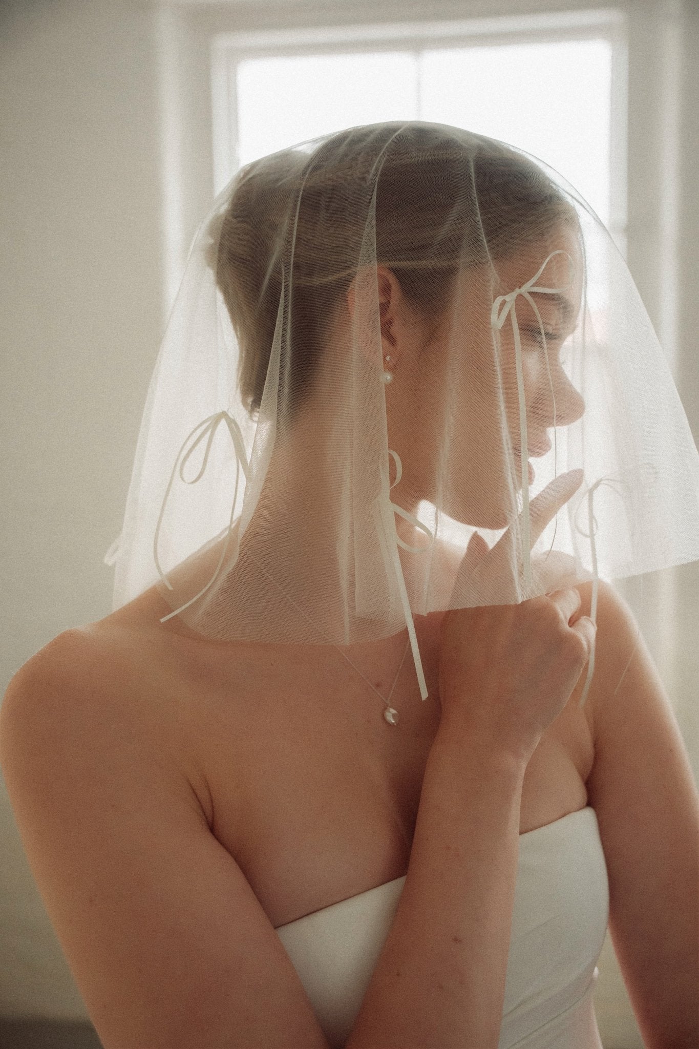Woman wearing a white wedding dress with a bow-embellished veil against a neutral background