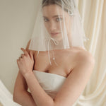 Woman wearing a white wedding dress with a bow-embellished veil against a neutral background