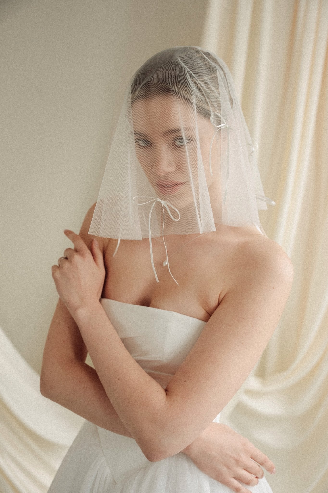Woman wearing a white wedding dress with a bow-embellished veil against a neutral background
