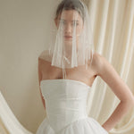 Woman wearing a white wedding dress with a bow-embellished veil against a neutral background