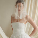 Woman wearing a white wedding dress with a bow-embellished veil against a neutral background