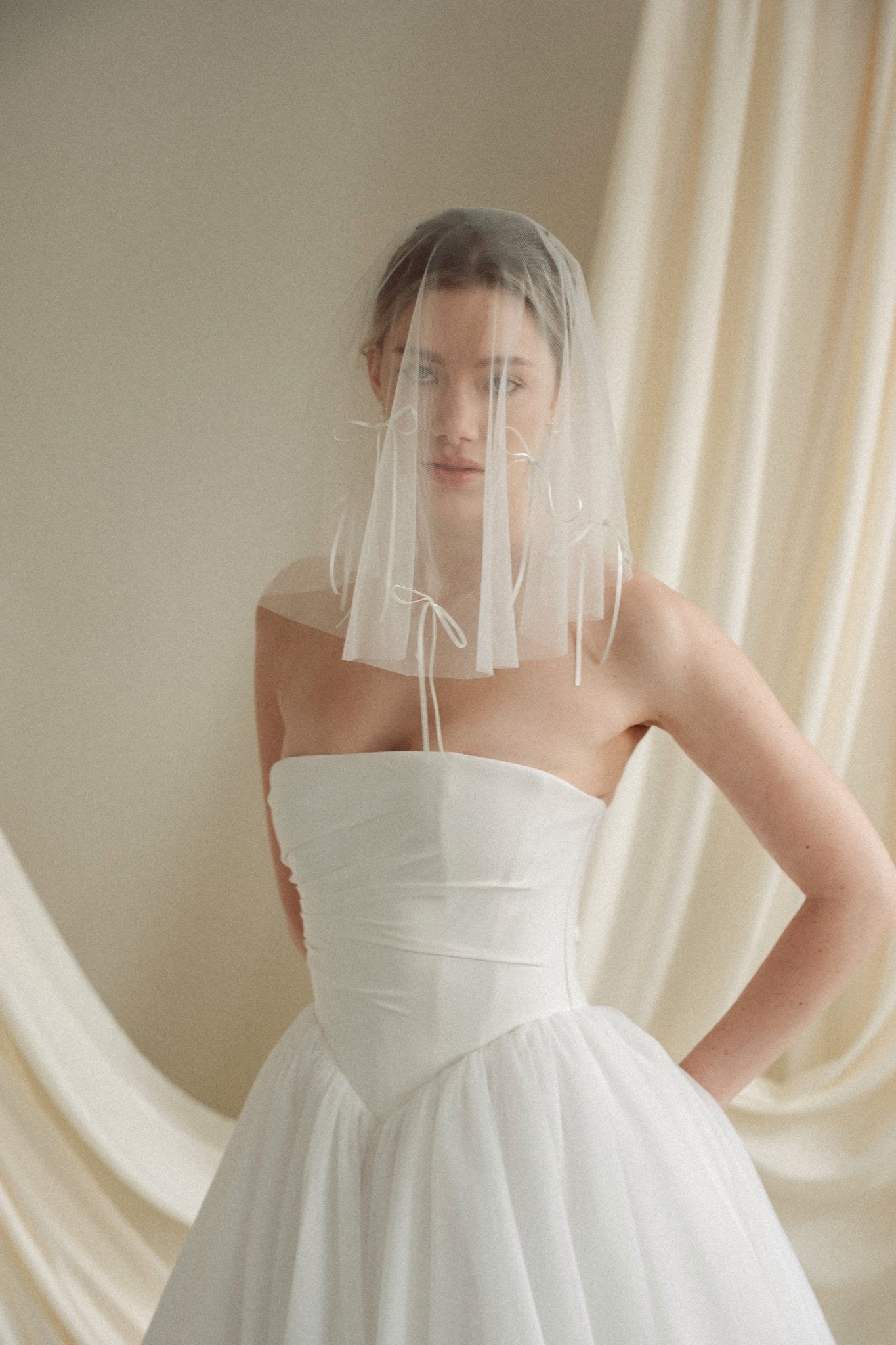 Woman wearing a white wedding dress with a bow-embellished veil against a neutral background