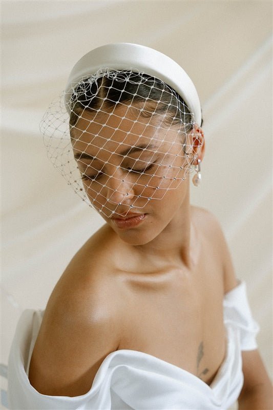 Merry Widow Graduated Crystal Birdcage Veil worn by a black bride with natural hair in a low bun with optional ivory satin padded headband and statement silver and pearl oblong hoop earrings