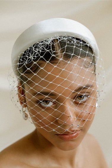 Merry Widow Graduated Crystal Birdcage Veil worn by a black bride with natural hair in a low bun with optional ivory satin padded headband and statement silver and pearl oblong hoop earrings