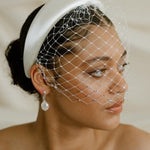 Merry Widow Graduated Crystal Birdcage Veil worn by a black bride with natural hair in a low bun with optional ivory satin padded headband and statement silver and pearl oblong hoop earrings