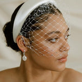 Merry Widow Graduated Crystal Birdcage Veil worn by a black bride with natural hair in a low bun with optional ivory satin padded headband and statement silver and pearl oblong hoop earrings