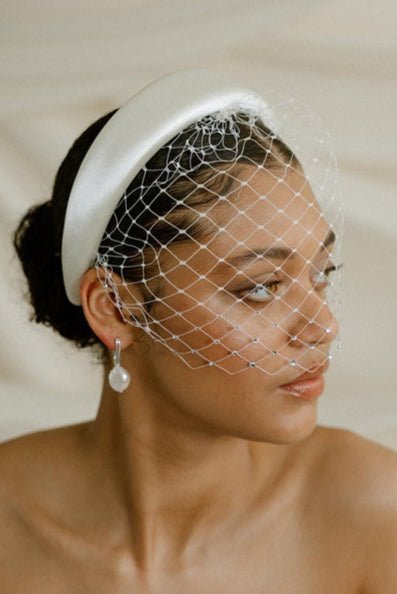 Merry Widow Graduated Crystal Birdcage Veil worn by a black bride with natural hair in a low bun with optional ivory satin padded headband and statement silver and pearl oblong hoop earrings