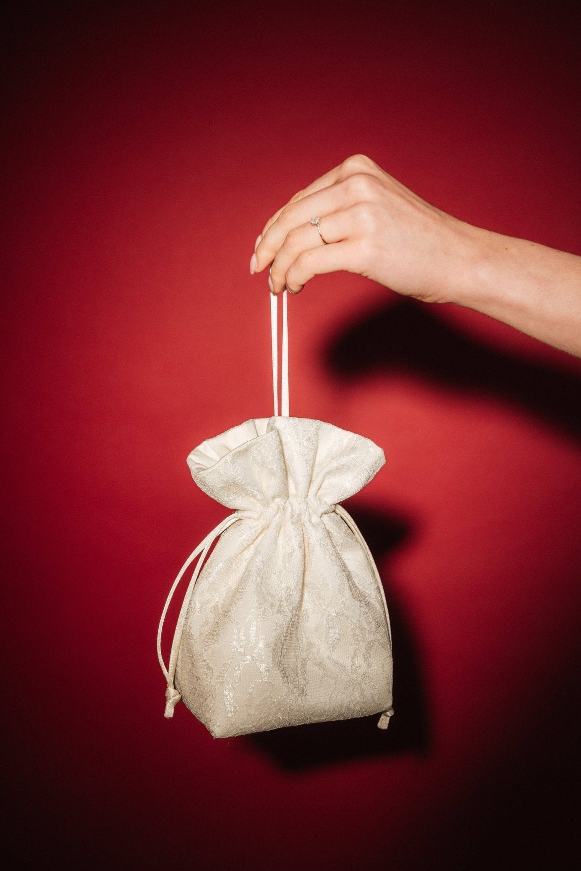 Hand holding a small ivory lace and satin drawstring bag against a red background