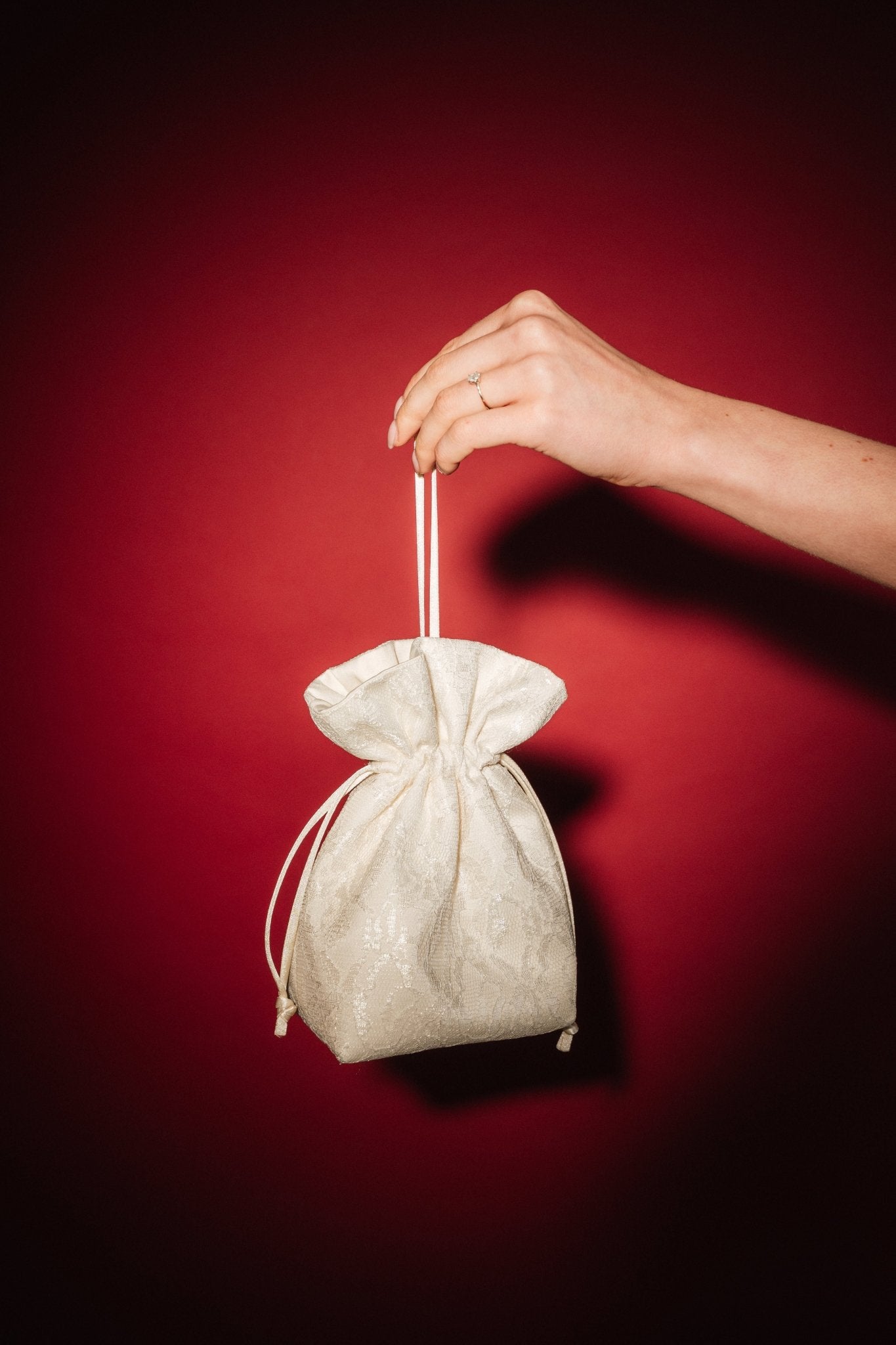 Hand holding a small ivory lace and satin drawstring bag against a red background