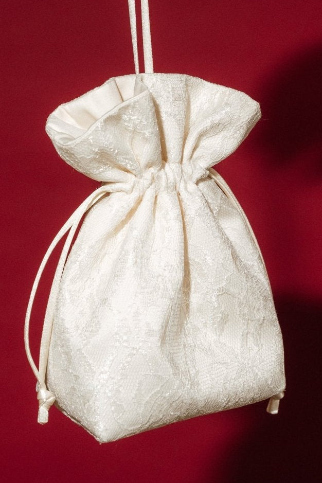 Hand holding a small ivory lace and satin drawstring bag against a red background