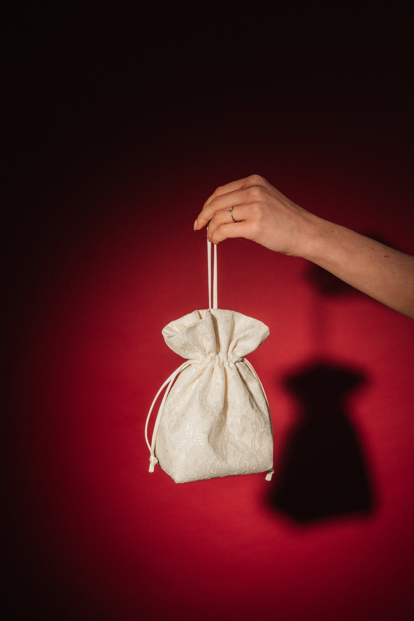Hand holding a small ivory lace and satin drawstring bag against a red background