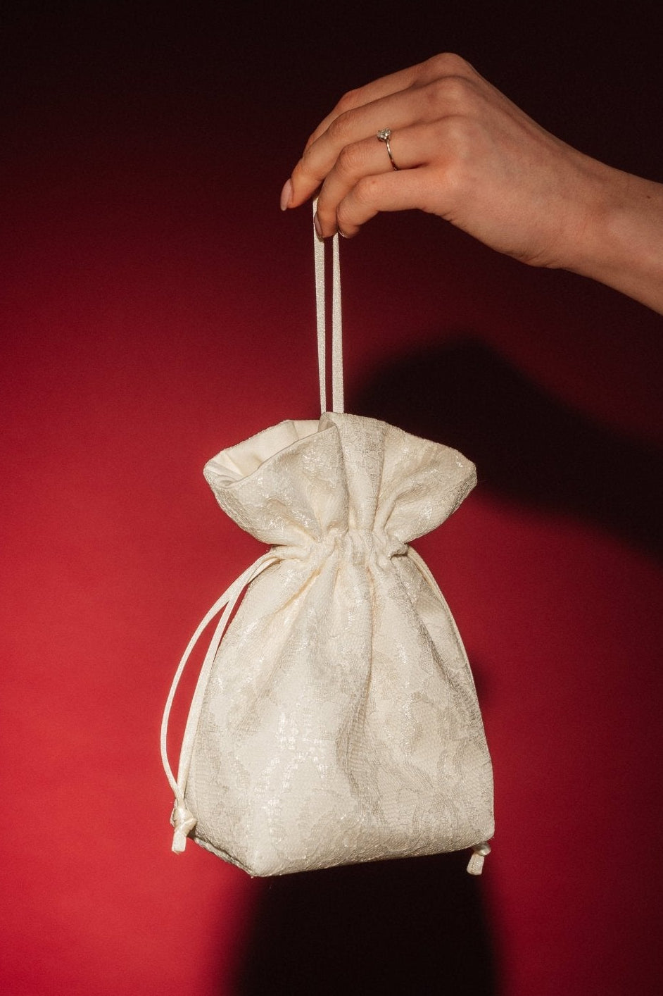 Hand holding a small ivory lace and satin drawstring bag against a red background