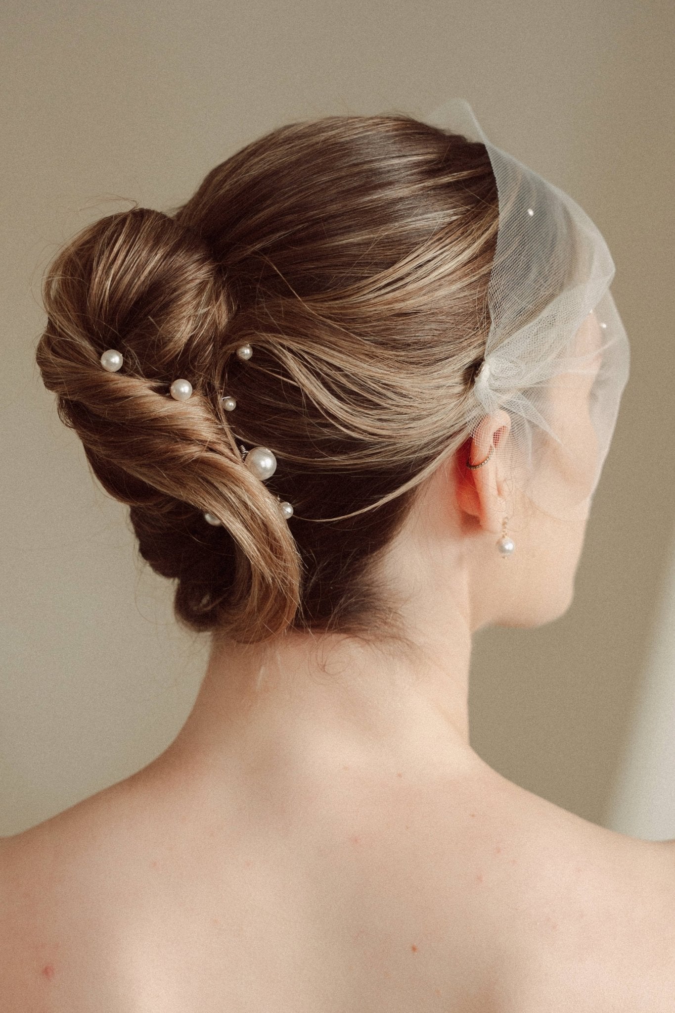 bride wears an ivory tulle mask style micro veil with small pearl embellishment with small pearl hair pins in a a soft chignon French twist updo and crystl and pearl stud drop earring