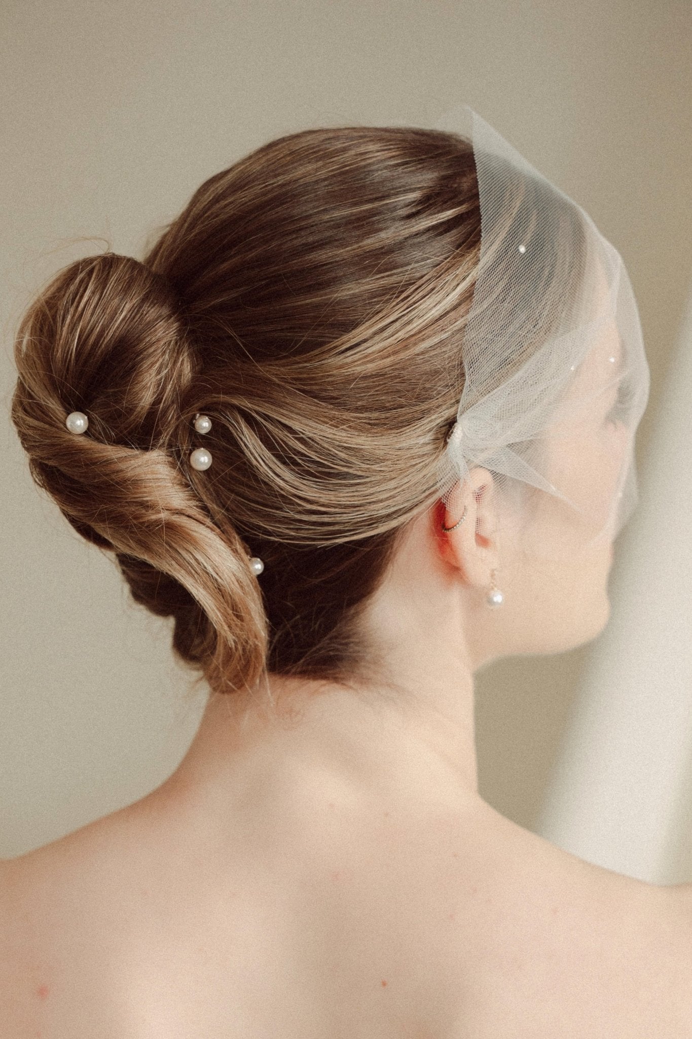 bride wears an ivory tulle mask style micro veil with small pearl embellishment with small pearl hair pins in a a soft chignon French twist updo and crystl and pearl stud drop earring