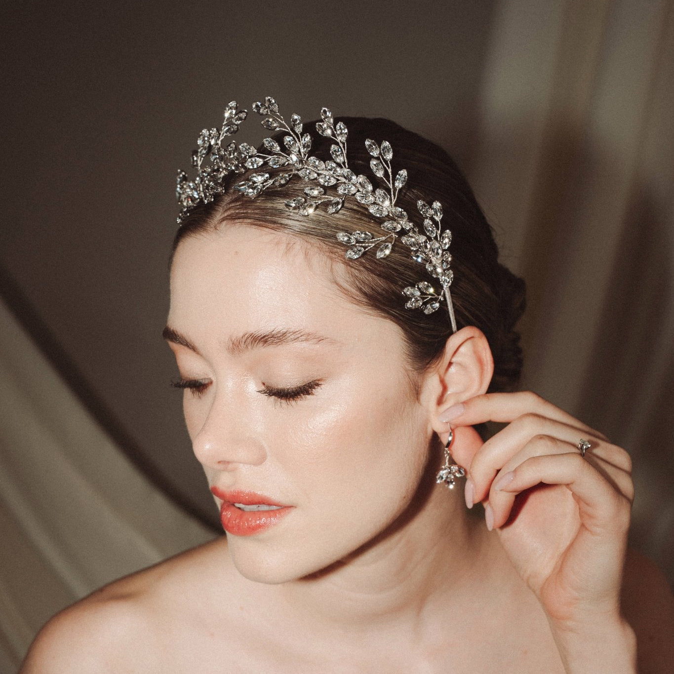 A bride wears a sparling silver crystal crown with hair pins positioned at it's base to create the effect of leaves growing out of the base of the headpiece she is adjusting her matching crystal drop earrings