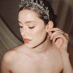 A bride wears a sparling silver crystal crown with hair pins positioned at it's base to create the effect of leaves growing out of the base of the headpiece she is adjusting her matching crystal drop earrings