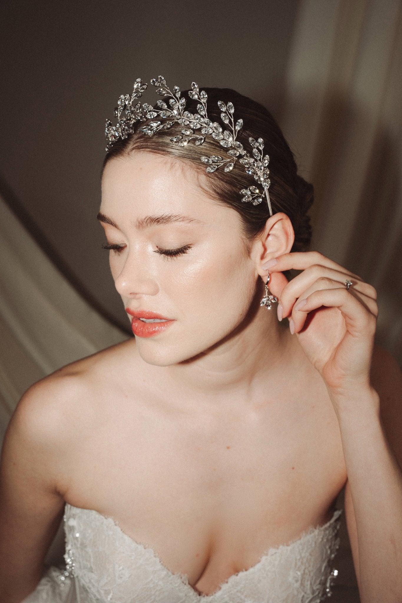 A bride wears a sparling silver crystal crown with hair pins positioned at it's base to create the effect of leaves growing out of the base of the headpiece she is adjusting her matching crystal drop earrings