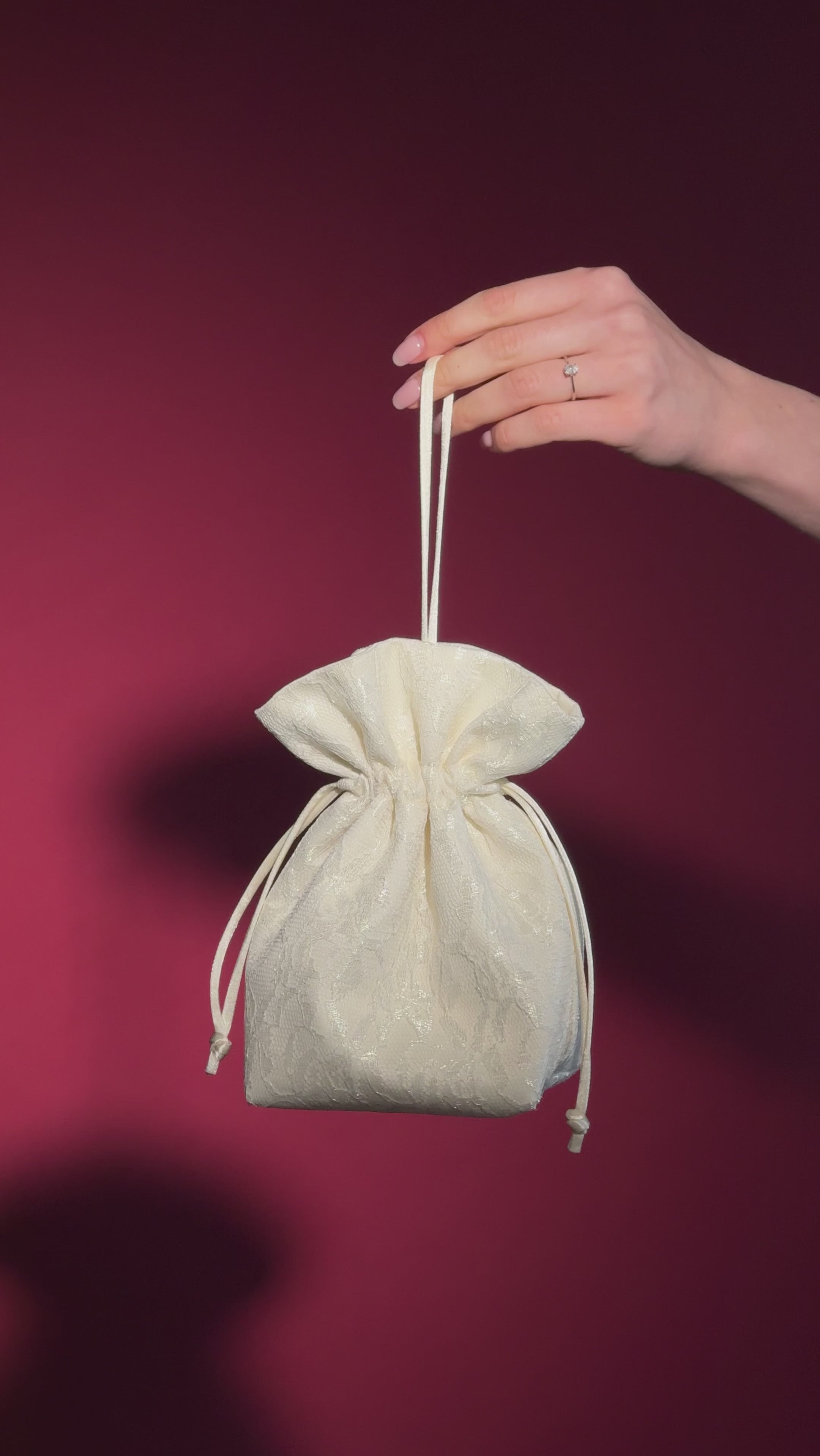 Hand holding a small ivory lace and satin drawstring bag against a red background